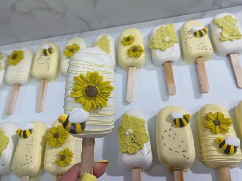 May include: A collection of decorative, bee-themed treats on wooden sticks. The treats are yellow and white, decorated with yellow flowers, honeycomb patterns, and small bee figures. The treats are arranged in rows.