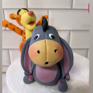 May include: A gray and purple figurine of Eeyore from Winnie the Pooh, with a black ear and a surprised expression. A figurine of Tigger is in the background.