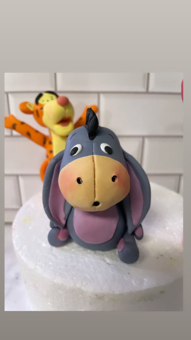 May include: A gray and purple figurine of Eeyore from Winnie the Pooh, with a black ear and a surprised expression. A figurine of Tigger is in the background.
