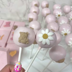 May include: Pink cake pops with white sticks, decorated with a teddy bear and daisy design. The cake pops are arranged on a white grid, with pink square treats in the background. The treats are perfect for a baby shower or birthday party.