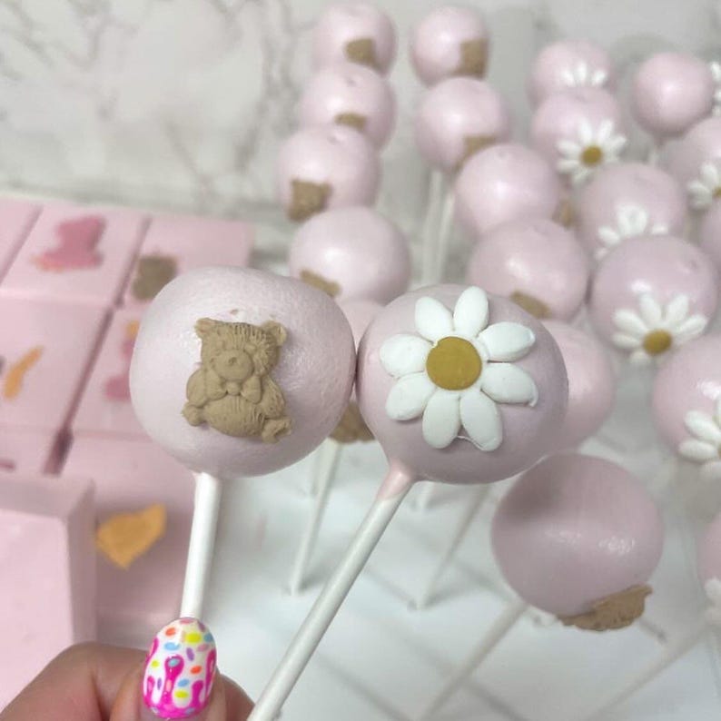 May include: Pink cake pops with white sticks, decorated with a teddy bear and daisy design. The cake pops are arranged on a white grid, with pink square treats in the background. The treats are perfect for a baby shower or birthday party.