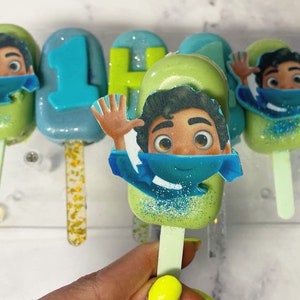 May include: A collection of colorful cake pops on sticks, decorated with cartoon character images and the number "1". The pops are in shades of green and blue, with glitter accents. The pops are arranged on a clear display.
