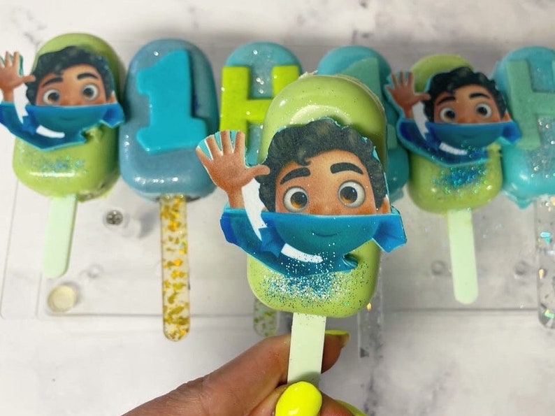 May include: A collection of colorful cake pops on sticks, decorated with cartoon character images and the number "1". The pops are in shades of green and blue, with glitter accents. The pops are arranged on a clear display.