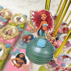 May include: Ariel-themed cake pops and decorated boxes. The cake pops are green with a blue rose and a character image. The boxes are pink and green, with character images and gold frames. Gold sticks are used to hold the cake pops.