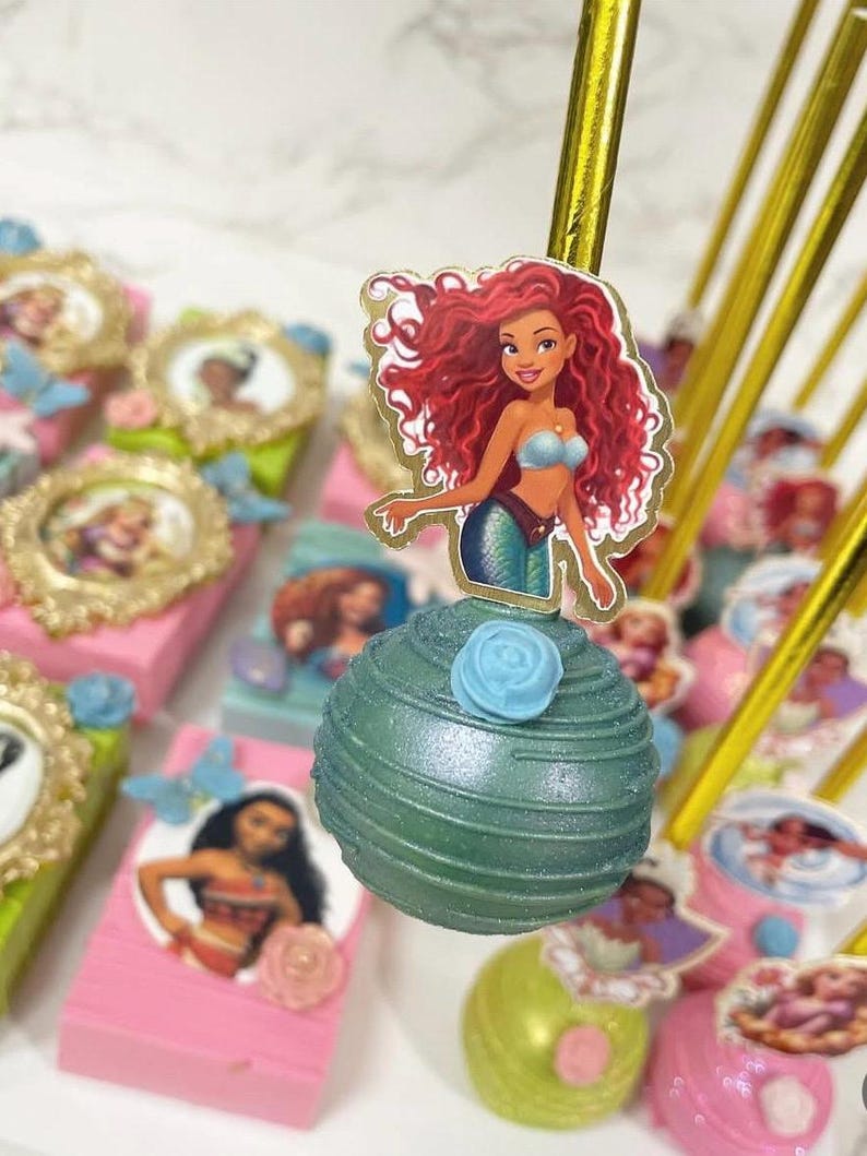 May include: Ariel-themed cake pops and decorated boxes. The cake pops are green with a blue rose and a character image. The boxes are pink and green, with character images and gold frames. Gold sticks are used to hold the cake pops.