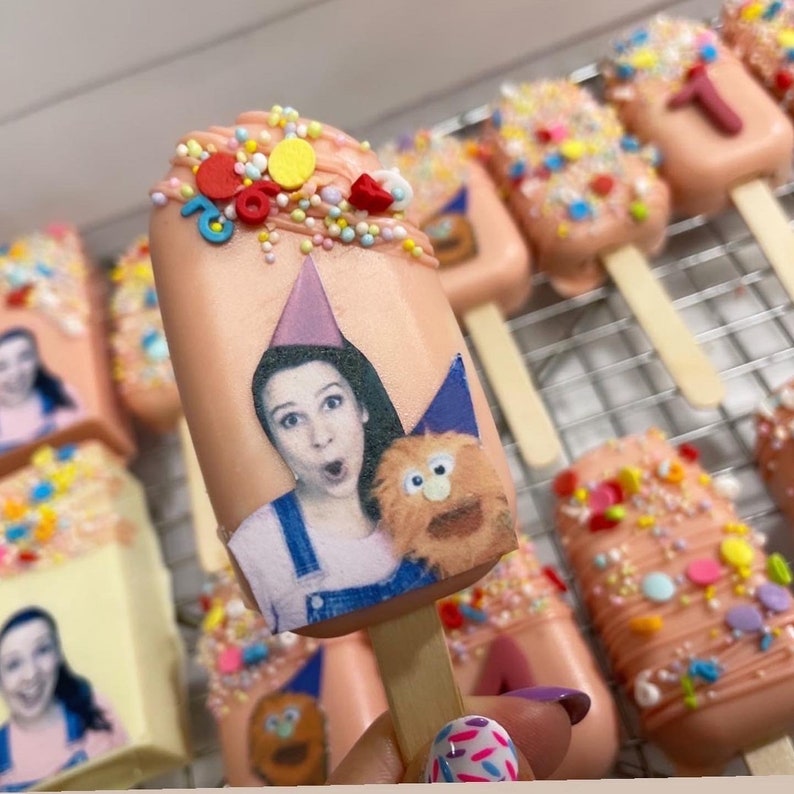 May include: Pink cake pops decorated with sprinkles and colorful candies. Each pop features a printed image of a person and a puppet, with a party hat. Some pops have the number "1" on them. The pops are on wooden sticks.