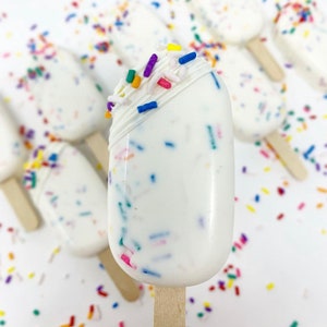 May include: White, oblong-shaped cake pops on wooden sticks, covered in white frosting and colorful sprinkles. Some pops have a white chocolate drizzle and are surrounded by more sprinkles. A sweet treat.