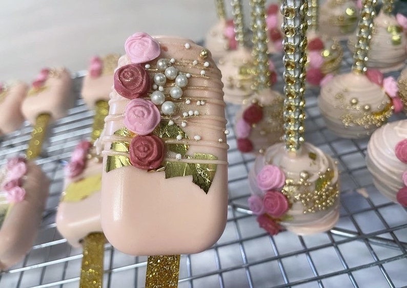 May include: Close-up of several decorated cake pops. The pops are light pink with gold accents, edible gold leaf, and small pink and burgundy roses. Some pops are on gold glitter sticks.