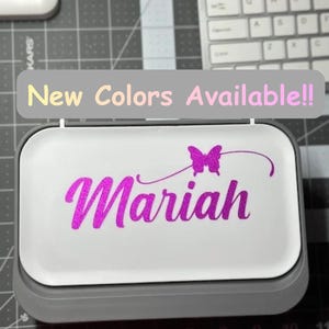 May include: A white and gray box with the name "Mariah" in bright pink script, accompanied by a butterfly graphic. The text "New Colors Available!!" is displayed above the box.