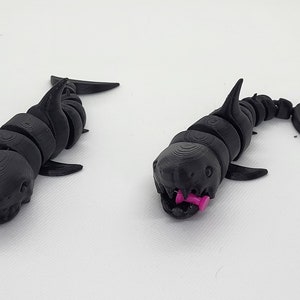 Può includere: Due giocattoli di squalo articolati neri. Uno squalo ha uno spillo rosa nella bocca.