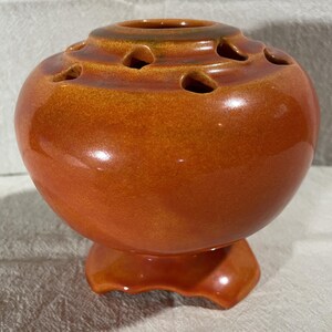 May include: A ceramic, orange-brown vase with a bulbous body and a flared base. The top of the vase has several evenly spaced holes. The vase has a glossy finish and is likely used for decorative purposes.