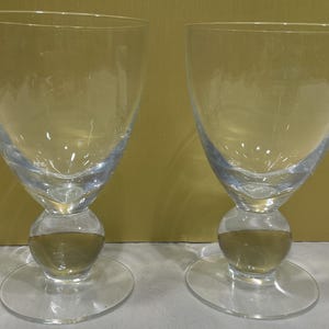May include: Two clear glass goblets featuring a wide, tapered bowl and a distinct spherical, ball-shaped stem resting on a flat, circular base. The glassware is transparent and smooth, showcasing a simple, functional design for serving beverages.