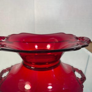 May include: A deep red glass bowl featuring a scalloped rim and two integrated side handles. The bowl has a wide, open top that tapers down to a smaller base, with a smooth, translucent finish that reflects light across its vibrant surface.