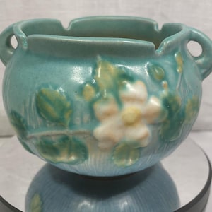May include: A blue glazed ceramic planter with two handles and a white floral design. The planter has a textured surface and is decorated with a white flower and green leaves.