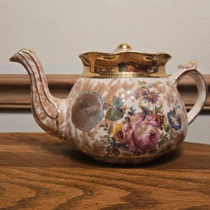 May include: A vintage teapot with a floral design featuring pink roses, purple and yellow flowers, and gold trim. The teapot has a light brown speckled background and a silver seal that reads "WEDGWOOD".