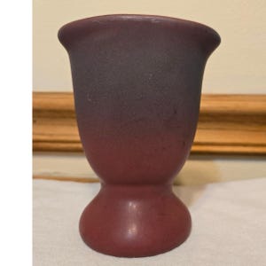 May include: A two-toned, burgundy-colored vase with a rounded base and a flared top. The vase has a smooth, matte finish and a gradient effect, transitioning from a darker shade at the top to a lighter shade at the base. The vase is approximately 5 inches tall.
