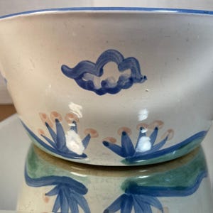 May include: A white ceramic bowl with a blue rim and painted blue cloud and floral designs. The bowl has a wide opening and a rounded shape. The floral designs have blue leaves and pink centers.