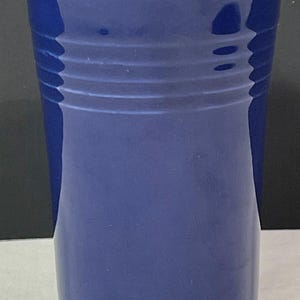 May include: A tall, cobalt blue ceramic drinking glass with a slightly flared rim and a smooth, glossy finish. The glass has a subtle, textured design around the middle.