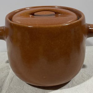 May include: A brown ceramic casserole dish with a lid. The dish has two handles and a small knob on the lid. The exterior is a glossy, speckled brown. A kitchenware item.