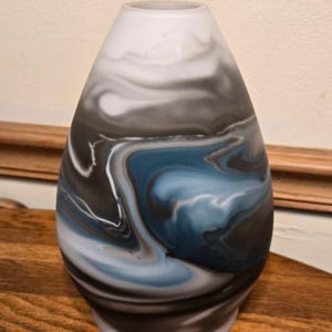 May include: A teardrop-shaped glass vase featuring a marbled pattern of swirling white, charcoal gray, and muted blue colors. The surface has a smooth, frosted finish with a tapered neck and a wider base, creating a fluid, organic appearance.