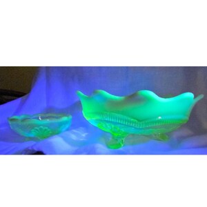 May include: Two pieces of green glass tableware. One is a small, round bowl with a scalloped edge. The other is a larger, footed bowl with a wavy rim and a textured design. Both pieces glow under a black light.