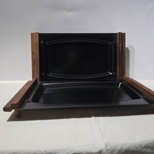 May include: A black serving tray with a rectangular shape, set within a wooden frame. The tray has a glossy finish and is open, revealing its interior. The wooden frame is a dark brown color.
