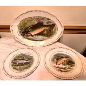 May include: Three oval porcelain plates with gold trim and painted images of fish. The largest plate features a largemouth bass, the medium plate features a herring, and the smallest plate features a trout.