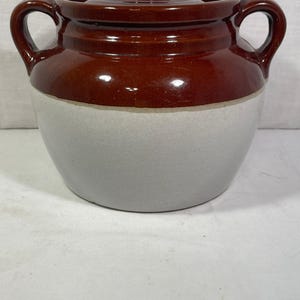 May include: A brown and white glazed ceramic bean pot with two handles and a lid. The pot is a traditional style with a rounded body and a wide mouth.