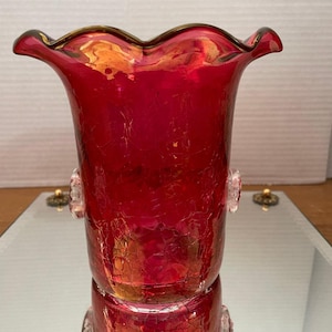 May include: A vibrant red glass vase with a crackle finish. The vase has a ruffled rim with a gold trim and two clear, decorative handles. The vase is reflected in the mirrored surface below.