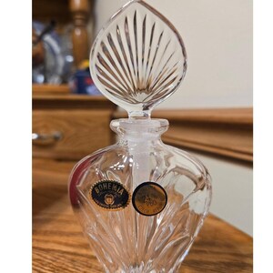 May include: A clear lead crystal perfume bottle with a fan-shaped stopper. The bottle features vertical fluted detailing and two circular stickers. One sticker reads Bohemia Czech Republic Lead Crystal Over 24%, and the other reads Bohemia 24% PbO.