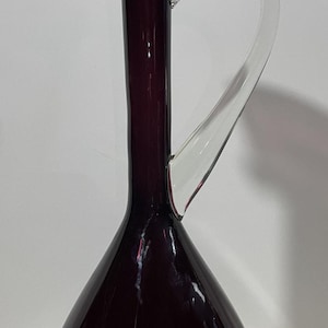 May include: A tall, elegant glass pitcher in a deep purple hue. It features a flared rim and a clear, arched handle. The pitcher's body has a rounded base that tapers to a slender neck.