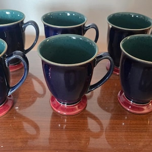 May include: A set of six ceramic mugs featuring a deep navy blue exterior and a speckled teal interior. Each mug has a rounded handle and sits on a distinct, flared red base with a speckled texture, creating a consistent look across the collection.
