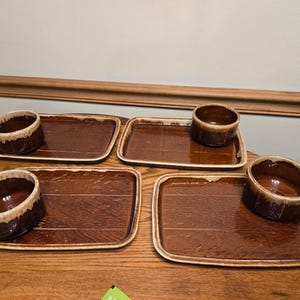May include: Four sets of brown ceramic serving dishes. Each set includes a rectangular tray and a small bowl. The bowls have a lighter brown rim. The trays have a wood-grain texture and a glossy finish. The sets are arranged on a wooden surface.