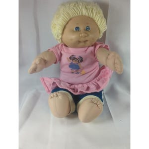 Pode incluir: Uma boneca de pele clara, olhos azuis e cabelo curto de lã loira. Veste uma t-shirt rosa de manga curta com o gráfico de uma figura com vestido azul, uma saia rosa com folhos e calções azul-escuros com detalhes brancos nas pernas.