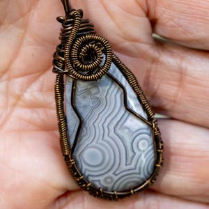 May include: A handcrafted pendant featuring a teardrop-shaped stone with gray and white swirling patterns. The stone is encased in a copper-colored wire wrap with a spiral design at the top. The pendant hangs from a delicate chain.