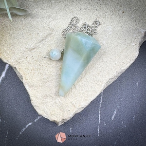 May include: A green and white crystal pendulum on a silver chain. The pendulum is shaped like a cone and is hanging from a white surface.