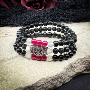 May include: A set of stacked black beaded bracelets featuring lava rock beads, white and magenta beads, and a silver-coloured centerpiece with a circular design. The bracelets are displayed on a light-coloured stone surface.