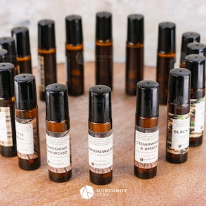 May include: A collection of 15 amber glass roller bottles with black caps and labels. The labels have different woodsy scents, including Cedarwood & Amber, Sandalwood, Mahogany, Black, and White Birch.