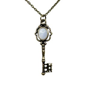 Può includere: Una collana con una chiave dorata con una pietra ovale bianca al centro. La chiave presenta dettagli intricati e un design decorativo.