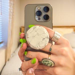 Puede incluir: Un soporte para teléfono con estampado de mármol blanco y gris sujeto a un teléfono inteligente gris. El teléfono se sostiene en una mano adornada con múltiples anillos, incluido un anillo grande de plata y un anillo con una piedra verde. Las uñas están pintadas de verde brillante.