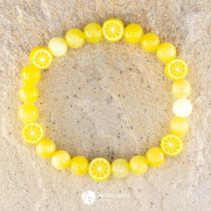 Puede incluir: Un brazalete de cuentas amarillas con dijes de rodajas de limón. El brazalete está sobre una superficie texturizada marrón claro.