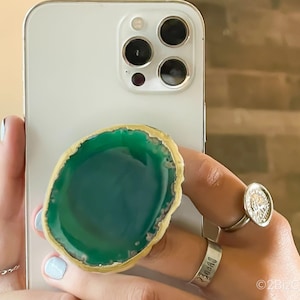 Könnte beinhalten: Ein grüner und goldener Achat-Handyhalter. Der Halter ist an einem weißen iPhone befestigt.