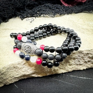 May include: Three beaded bracelets in a stack. The bracelets feature black, red, white, and navy blue beads. A silver-coloured cylindrical bead with a compass design is centered on one bracelet. The bracelets are displayed on a stone surface.