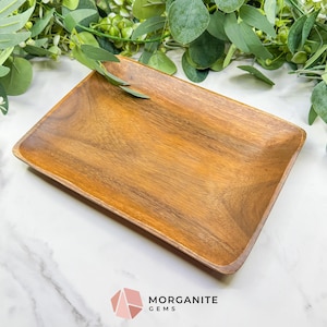 Puede incluir: Una bandeja rectangular de madera con un acabado marrón cálido. La bandeja tiene esquinas ligeramente redondeadas y una superficie lisa. Se ve follaje verde en el fondo. El logotipo de Morganite Gems está en la esquina inferior derecha.
