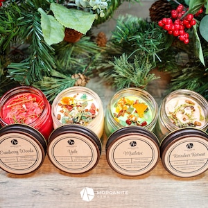 Op de afbeelding: Vier glazen potkaarsen met deksels, elk met een andere geur en kristallencombinatie. De geuren zijn Cranberry Woods, Yule, Mistletoe en Reindeer Retreat. De kaarsen zijn omringd door groen en rode bessen.