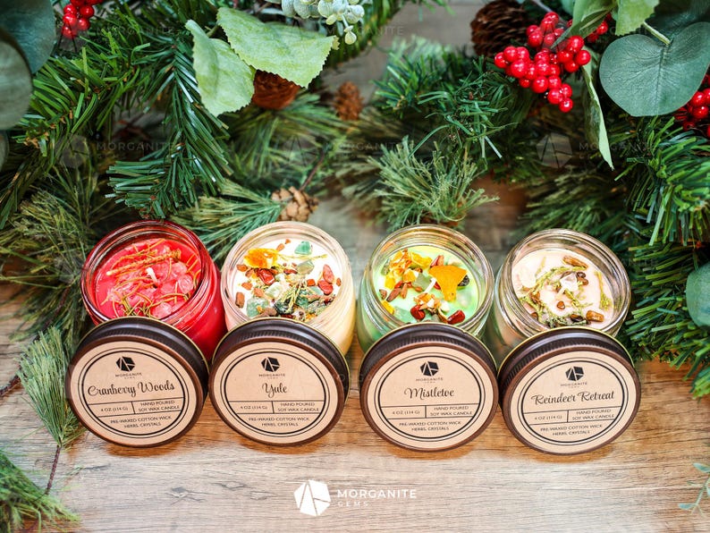 Op de afbeelding: Vier glazen potkaarsen met deksels, elk met een andere geur en kristallencombinatie. De geuren zijn Cranberry Woods, Yule, Mistletoe en Reindeer Retreat. De kaarsen zijn omringd door groen en rode bessen.