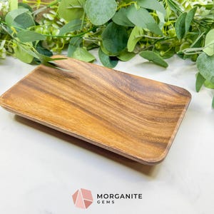 May include: A rectangular wooden tray with rounded corners. The tray is made of a light brown wood with a visible grain pattern. The tray is empty and sits on a white surface, with green foliage in the background. The logo for Morganite Gems is in the bottom right corner.