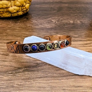May include: Copper cuff bracelet with seven colorful gemstones. The bracelet is resting on a white crystal against a wooden background. The gemstones are black, blue, black, green, yellow, red, and black.
