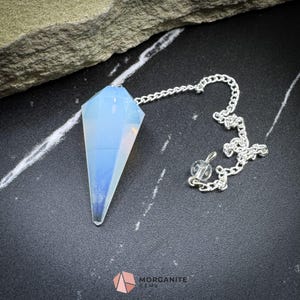 Puede incluir: Un péndulo de ópalo blanco con una cadena de plata. El péndulo tiene forma de lágrima puntiaguda y está colgado sobre una superficie negra.