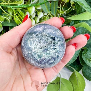 May include: A polished, spherical gemstone with a mix of blue, green, and white hues. The stone is held in a hand with red nail polish, surrounded by green foliage. The logo "MORGANITE GEMS" is visible.
